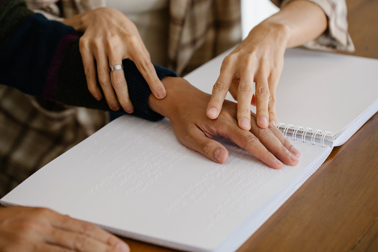 A caregiver guides a person with Braille reading using a tactile method and supportive touch.